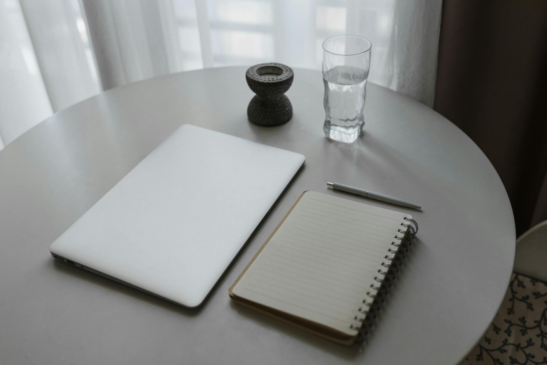 A serene minimalist workspace with a laptop, notebook, and glass of water on a round table.