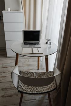 A clean and stylish home office desk with a laptop, notebook, and glass of water by a window.