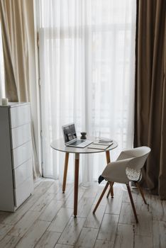 A minimalist workspace featuring a round table, chair, and laptop in a sunlit room with large curtains.