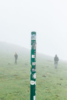 雾气缭绕的风景中，一根贴有贴纸的杆子立在背景里，两个人在徒步旅行