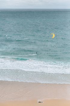 A lone kitesurfer glides over the ocean water near a sandy beach, captured from above.