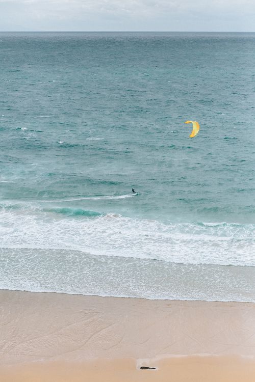 Free A lone kitesurfer glides over the ocean water near a sandy beach, captured from above. Stock Photo