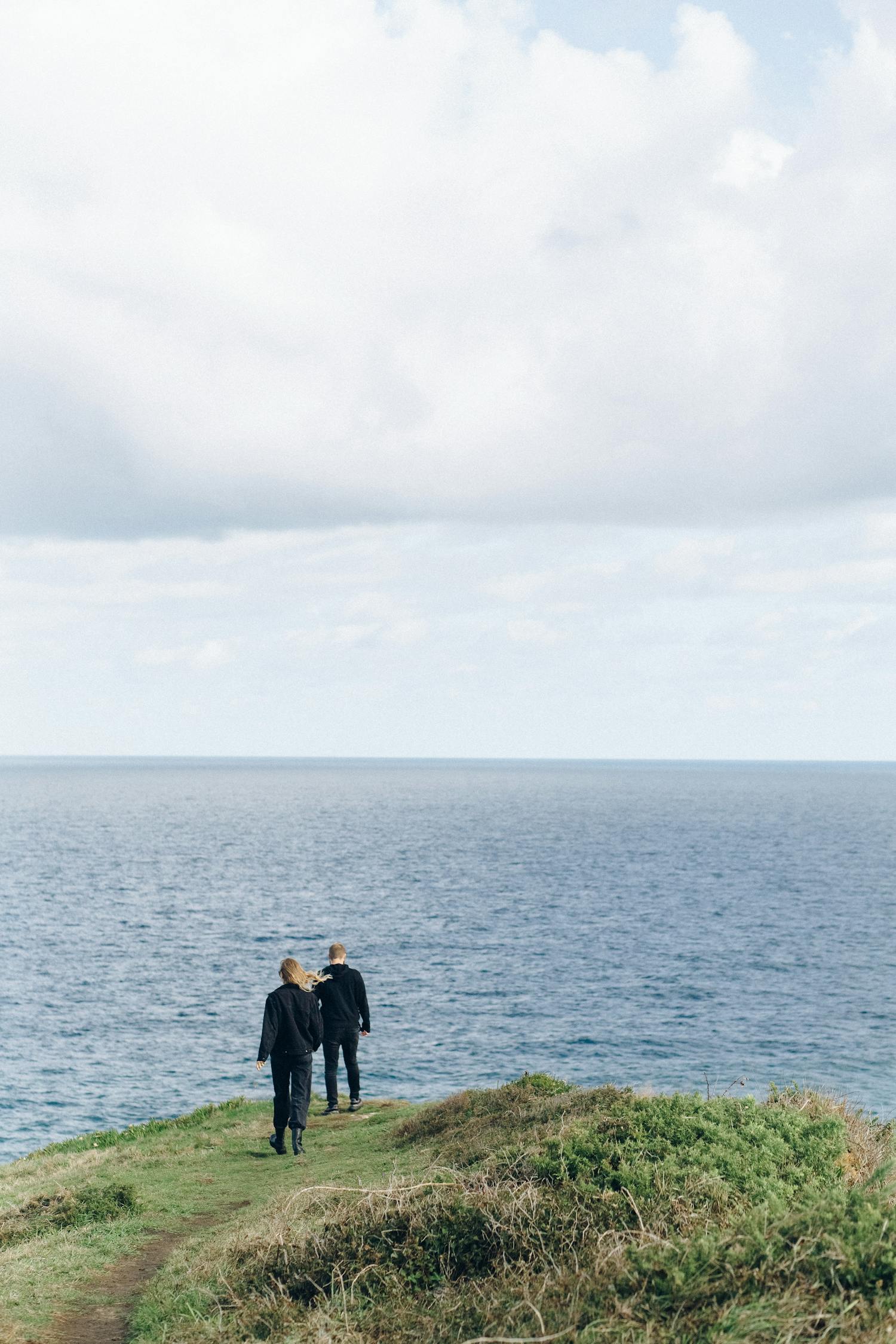 Couple enjoying a peaceful walk along a grassy cliff overlooking the ocean under a cloudy sky.
