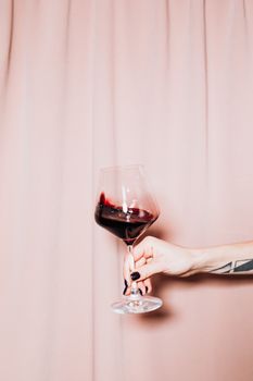 一只手优雅地握着红酒杯，置于柔软的粉红色窗帘前，流露出放松之感