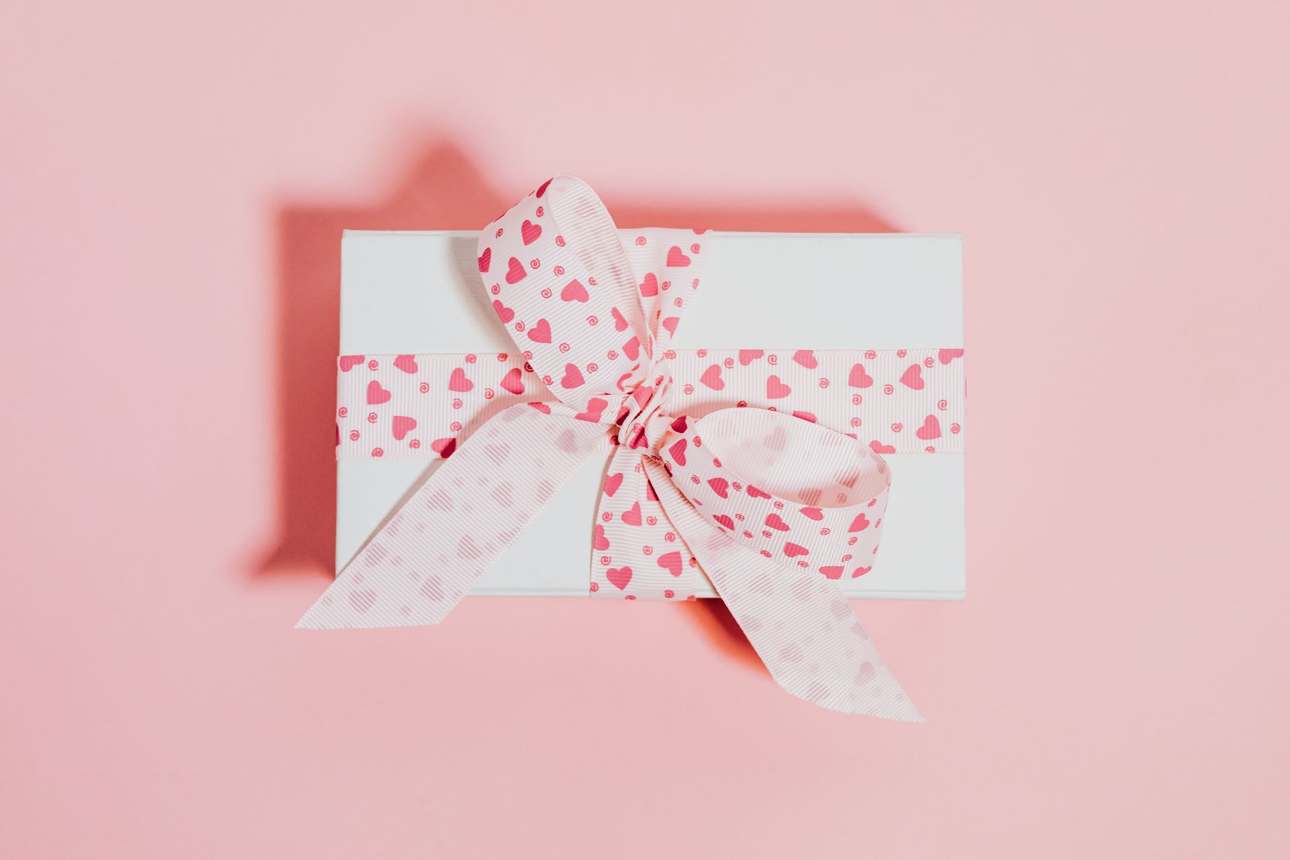 White gift box with pink heart-patterned ribbon on a soft pink background, ideal for celebrations.