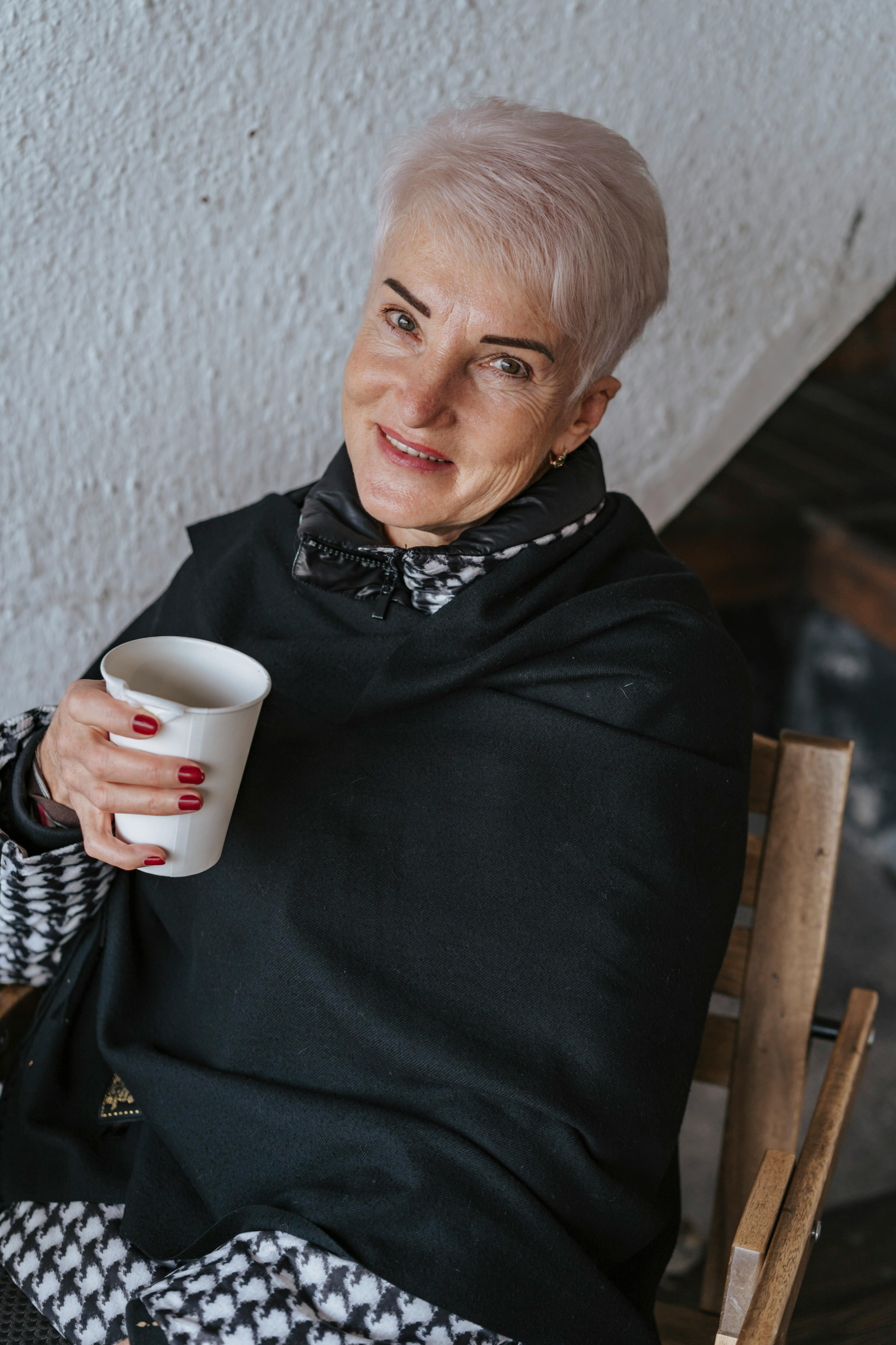 一位开朗的短发年长女性坐在室内，喝着咖啡，笑容温暖