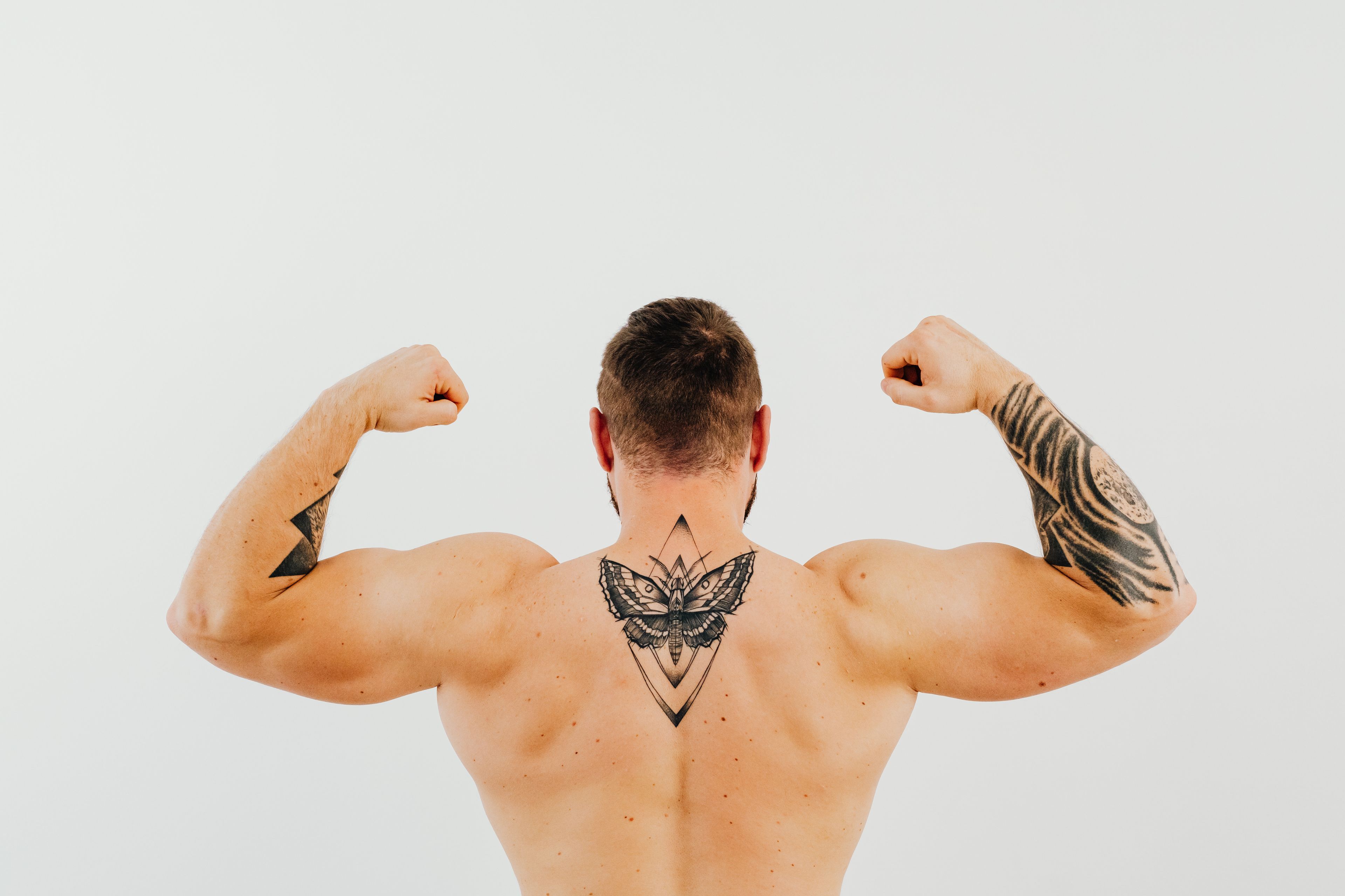 Shirtless man showcasing tattoos and biceps while flexing in a studio setting.