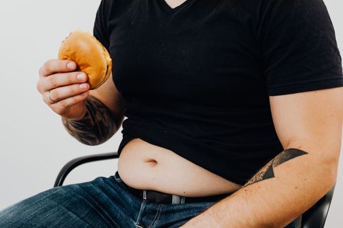 Free Close-up of a man holding a burger, showcasing his torso and tattooed arm. Casual and relaxed setting. Stock Photo