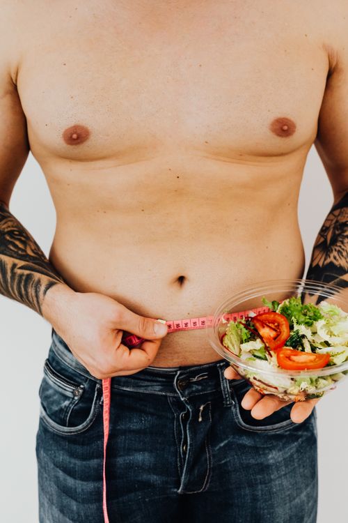 Free Tattooed man measuring waist while holding a salad bowl for healthy lifestyle and weight loss concept. Stock Photo
