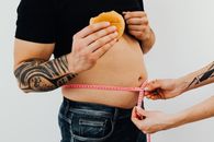 一名纹身男子拿着汉堡，另一个人正用卷尺测量他的腰围