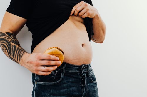 Free Man with tattoos holding a cheeseburger against his bare belly, wearing jeans and a black shirt. Stock Photo