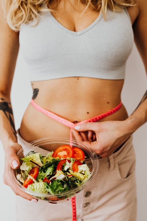 Free Fit woman in a crop top holds a salad bowl while measuring her waist, emphasizing healthy lifestyle and weight loss. Stock Photo
