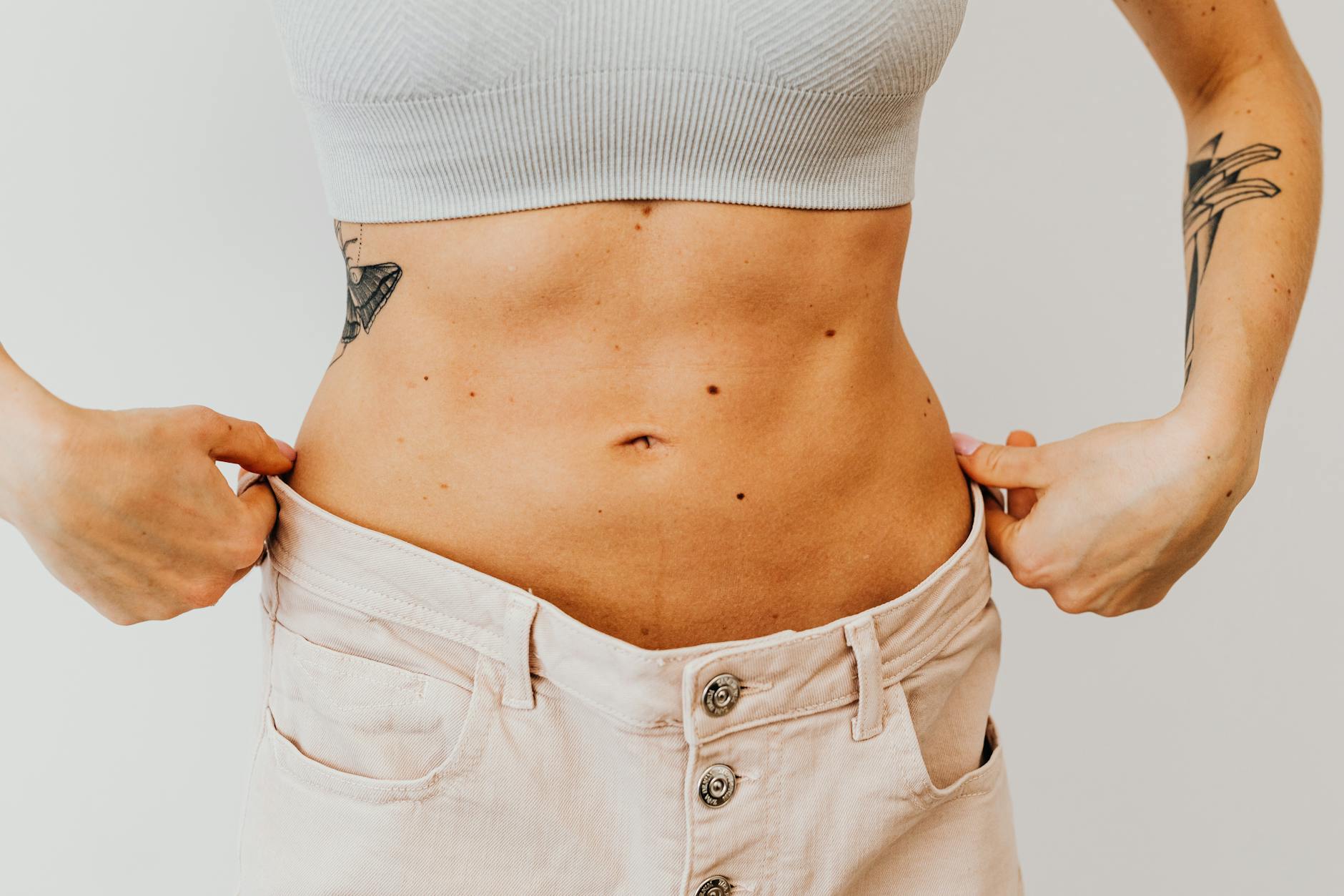 A woman shows weight loss progress by holding loose pants around her waist, highlighting tattoos.