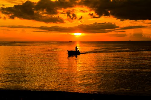 Free A stunning sunset with a silhouetted boat on a tranquil sea reflecting vibrant orange hues. Stock Photo