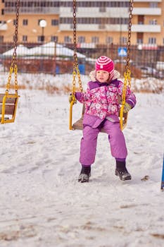 Young child in warm clothing enjoying a swing in a snowy playground during winter.