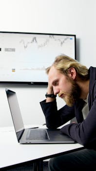 Frustrated man with beard at laptop, financial chart on screen.
