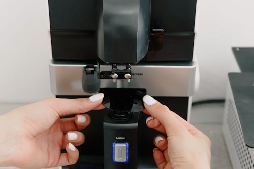 Unrecognizable female with manicure adjusting automatic dispenser while making aromatic hot drink on modern black coffee machine placed near wall