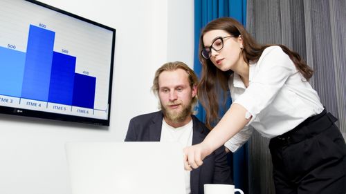 Free Two colleagues analyzing business data on a laptop with a presentation screen in the background. Stock Photo