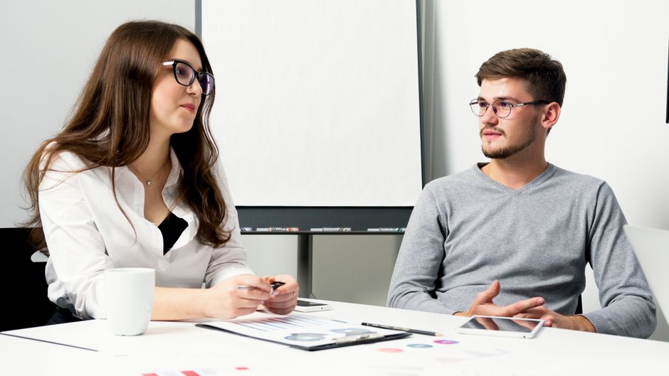 Two professionals in an office environment discussing work over a desk with documents.