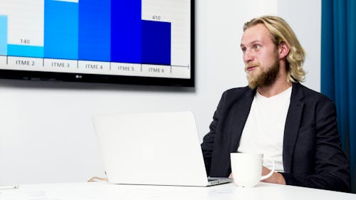 A Man in Black Suit Jacket Sitting at a Desk with a Laptop
Beside a Mug