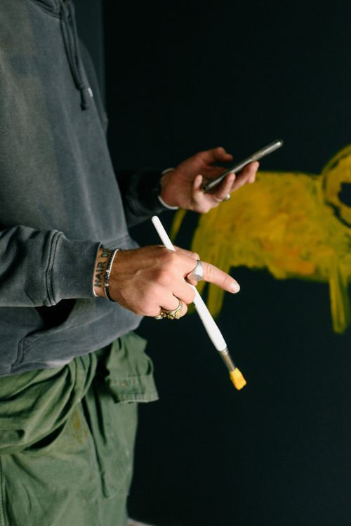 Free Side view of anonymous male painter with paintbrush in hand surfing cellphone while standing near black wall with painting in studio Stock Photo