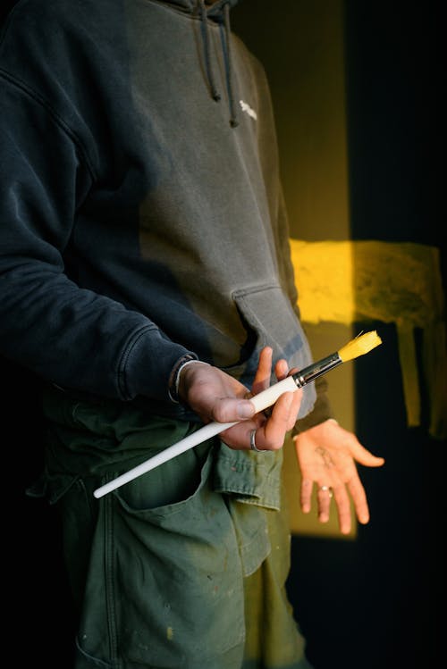 Unrecognizable male artist in uniform standing with paintbrush near black wall with yellow painting in bright sunlight while creating artwork