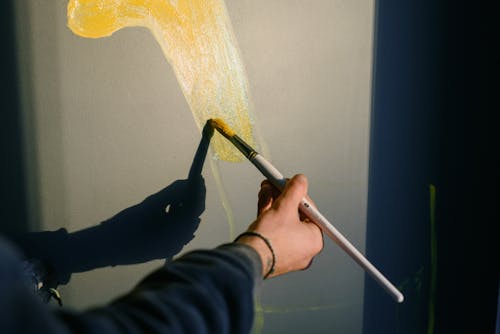 Unrecognizable professional male painter drawing yellow gouache with paintbrush in sunlight while casting shadow on black wall while creating artwork
