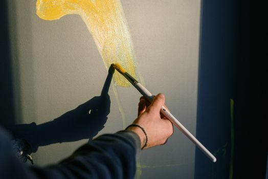 职业男画家在阳光下用画笔绘制黄色水粉画，创作艺术品时，他的身影在黑墙上投下阴影，面目已非