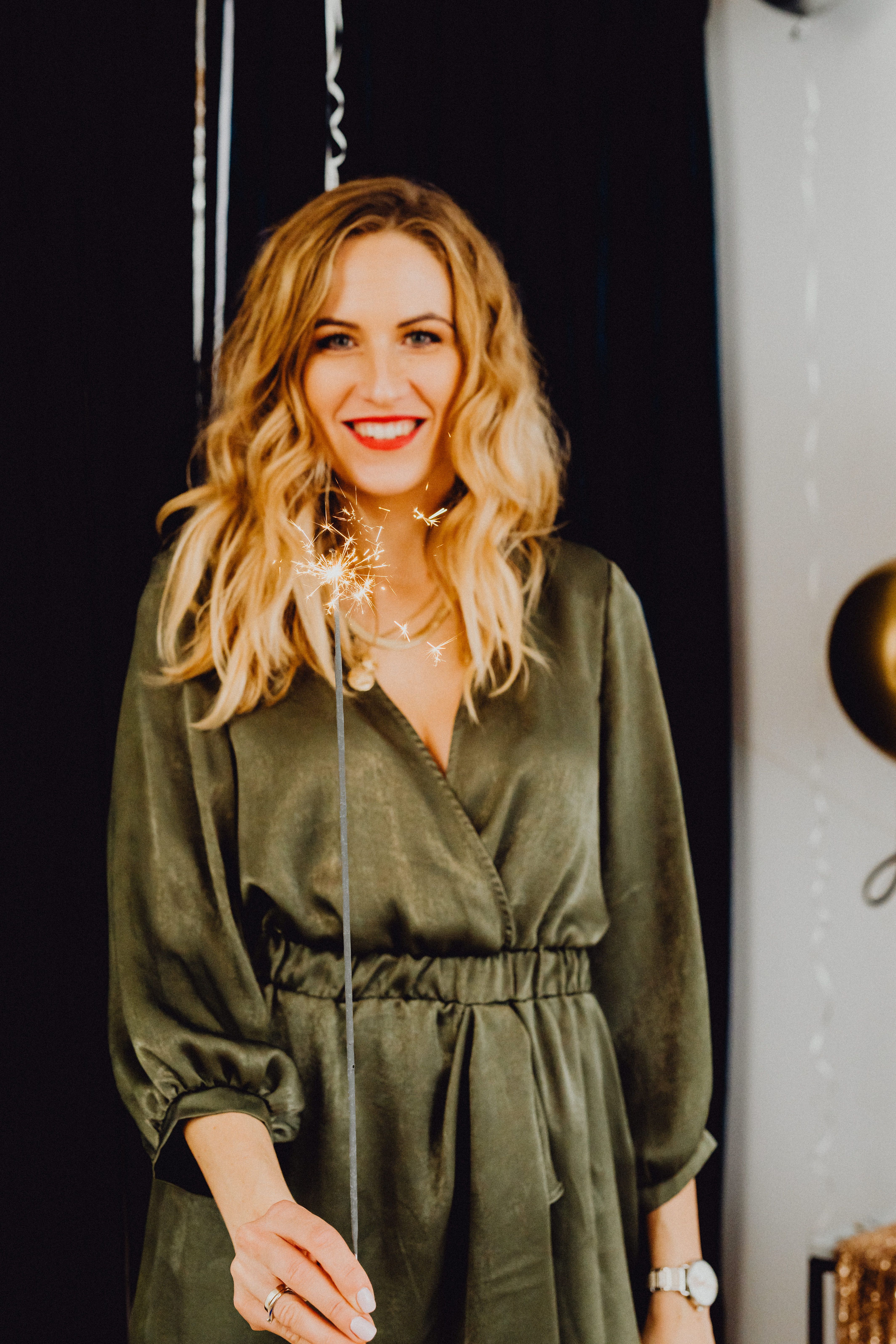 Smiling woman with blonde hair holding sparkler indoors, celebrating a joyful occasion in style.