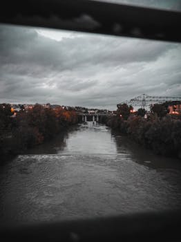 View from fence of ridge on river between banks covered with plants under gloomy cloudy sky