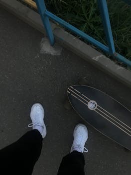 From above of crop unrecognizable person in activewear and sneakers standing on asphalt road near skateboard