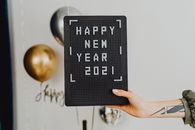 他手里拿着一块写着“2021年新年快乐”的信板，背景装饰着气球