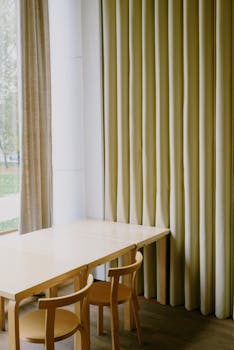 A cozy minimalist dining area with wooden chairs, table, and large window draped with curtains.