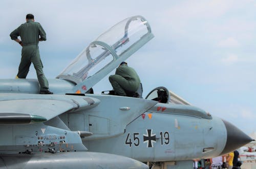 Low angle of unrecognizable male pilots in uniforms standing on aged gray multirole combat aircraft before flight against cloudy sunset sky