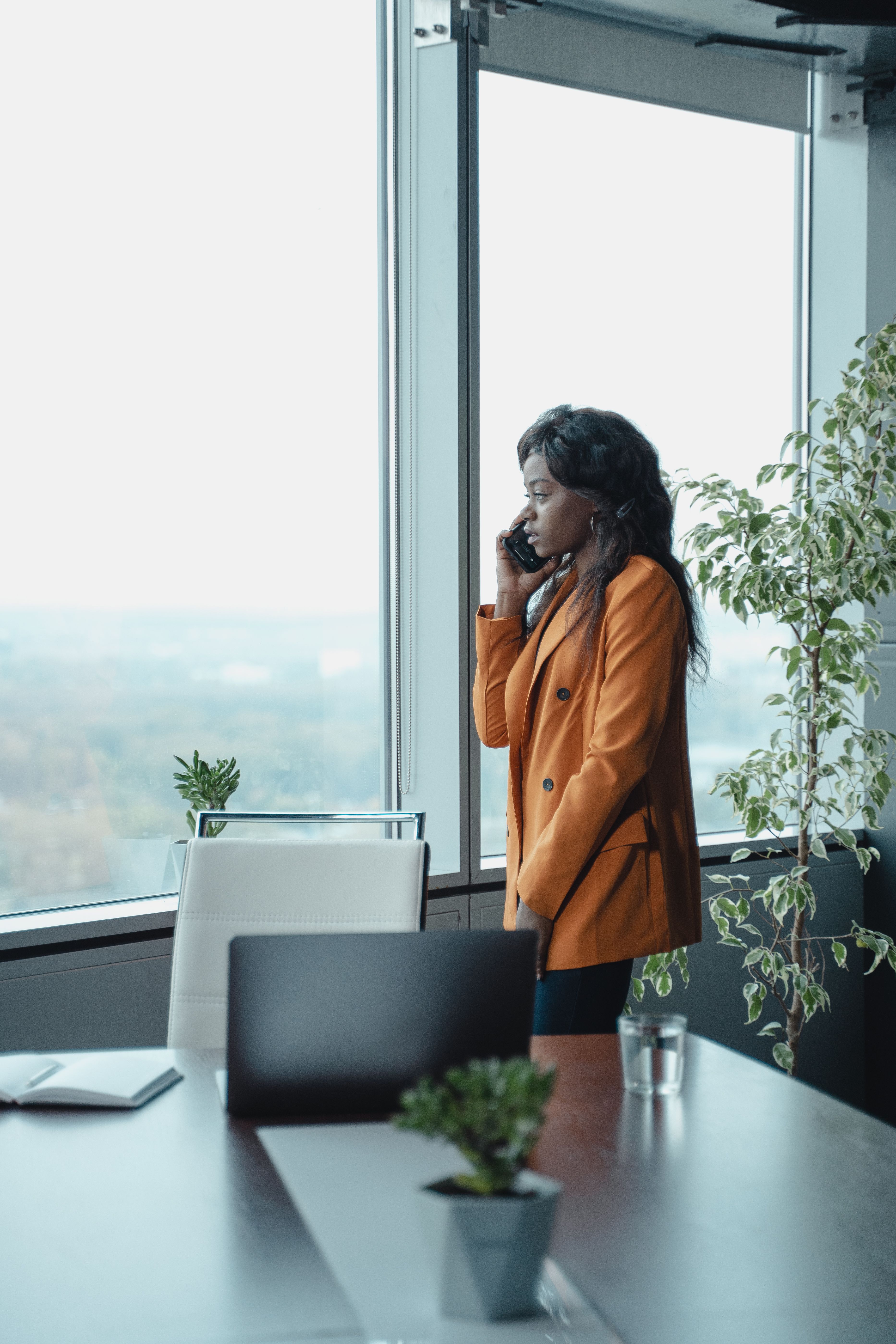 一位自信的女人穿着橙色夹克，在大办公室的窗边打电话