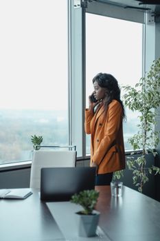 一位自信的女人穿着橙色夹克，在大办公室的窗边打电话