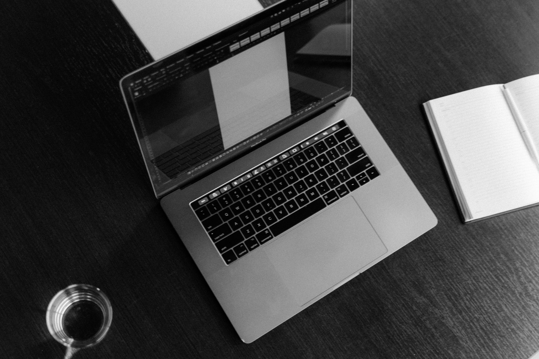 A high-angle black and white shot of a laptop, notebook, and glass of water on a wooden table.