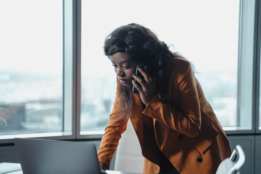 办公室里，一位穿着橙色外套的女商人正在使用智能手机和笔记本电脑