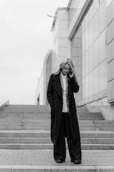 A professional woman in an overcoat talks on the phone while walking down concrete steps in the city.