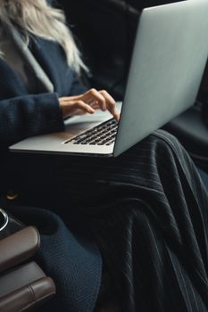 Woman in a blue coat typing on a laptop while sitting in an office setting.