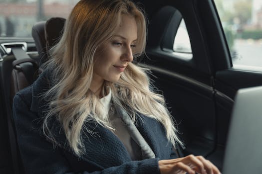 Blonde woman working on laptop in luxury car backseat, focused on her task.