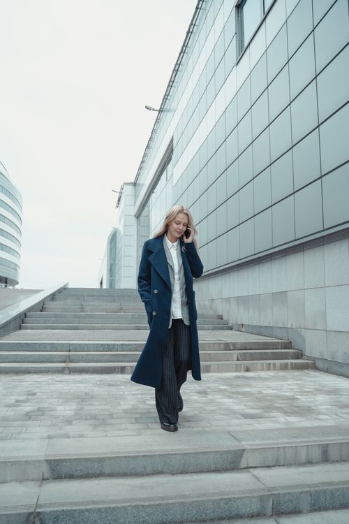 Free Confident woman in blue coat walking down outdoor stairs, talking on phone, showcasing urban lifestyle. Stock Photo