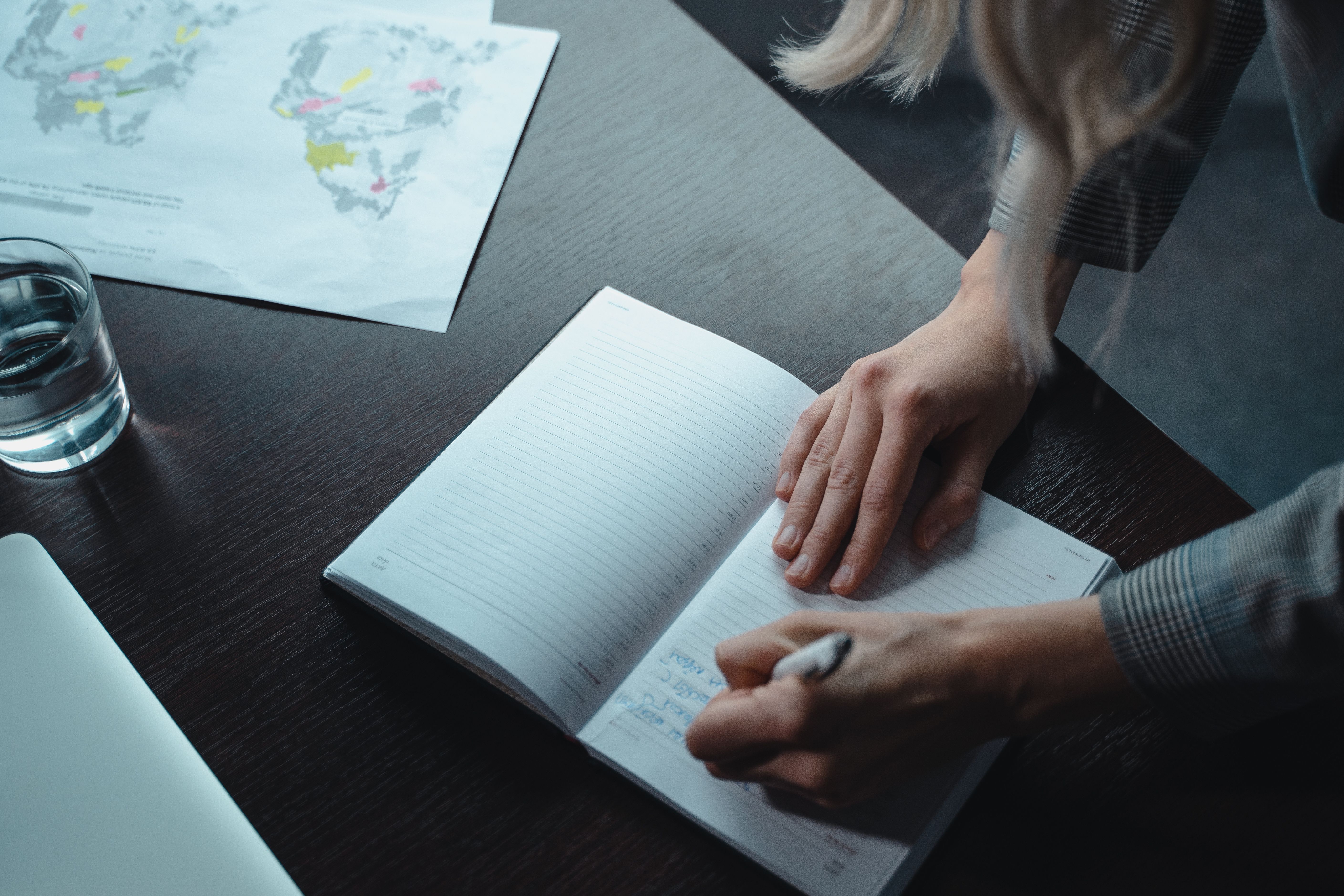 Woman writing in a notebook at a wooden table with a map and laptop nearby.