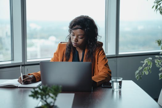 A professional woman in an orange blazer working on a laptop in a bright, modern office setting.