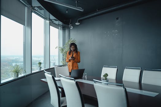 Elegant woman in orange blazer using smartphone in a stylish conference room with city view.
