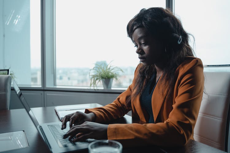 A Woman In An Orange Blazer Typing On A Laptop