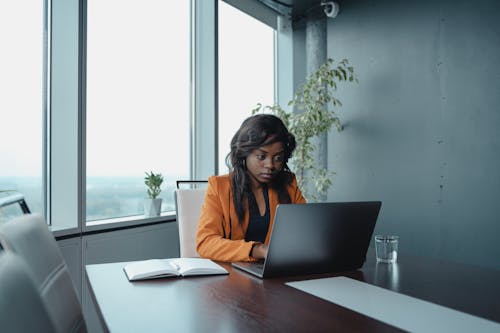 Woman in Orange Blazer Sitting at the Table Using a Macbook