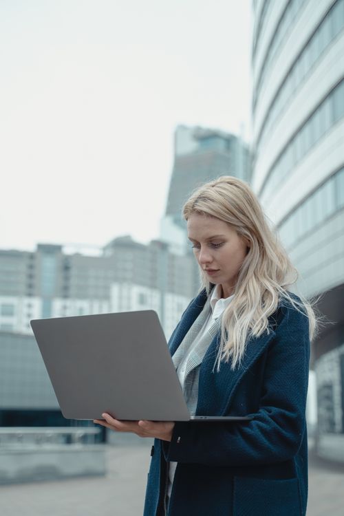 Free Caucasian woman with blonde hair using laptop outside modern office buildings. Stock Photo