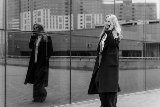 Monochrome image of a businesswoman in a coat walking while talking on her phone, city reflection.