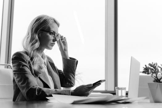 Black and white photo of a businesswoman working with a phone and laptop in a modern office setting.